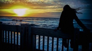 Silhouette of young woman sitting on sea front fence at dawn